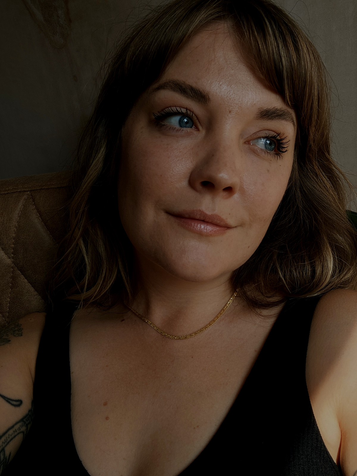 Close up of Lyzi sitting on a vintage chair in front of a plaster wall, wearing simple black underwear and a Daisy x Estee Lalonde snake chain.