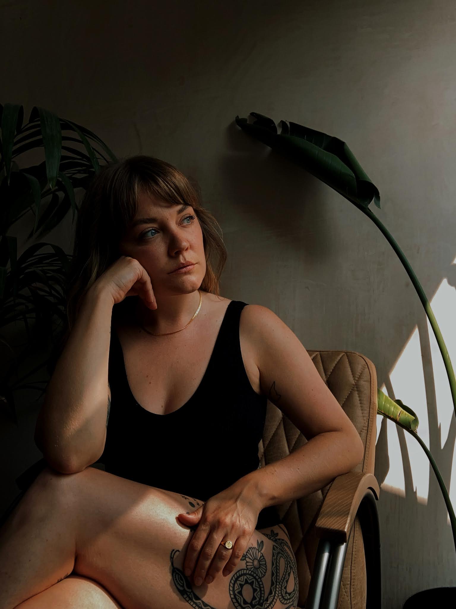 Wearing Daisy London snake chain Lyzi sitting on a vintage chair in front of a plaster wall and large plants. Wearing simple black underwear and a Daisy x Estee Lalonde snake chain and forget me not signet ring.