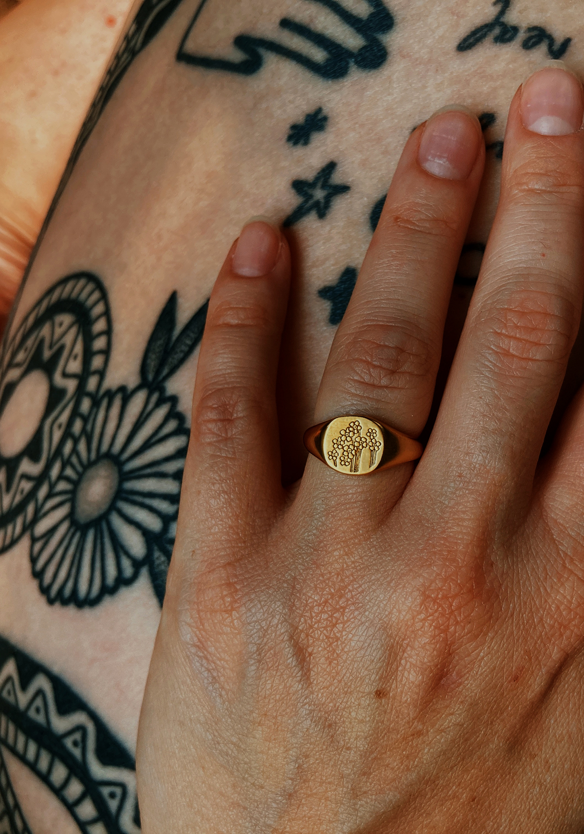 Close up of hand wearing Daisy forget me not signet ring, with tattooed legs in the background